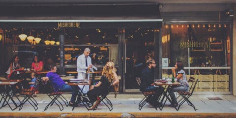 Outdoor dining scene at Mishiguene restaurant with diners sitting at tables and a waiter attending to guests.