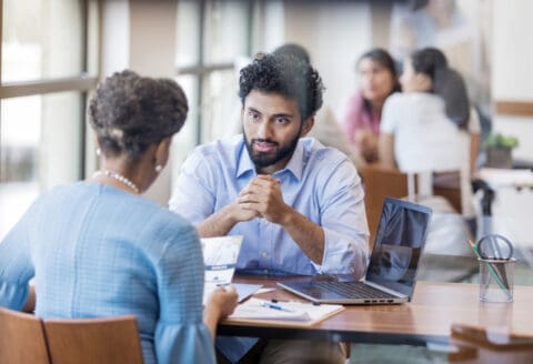 A man and woman having a conversation at a table with a laptop and documents in a professional setting.