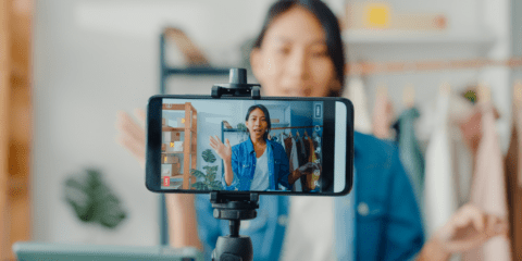 Person filming themselves with a smartphone in front of clothing rack.