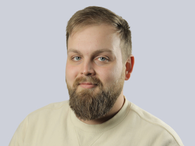 Bearded man in a beige sweater smiling against a light gray background.