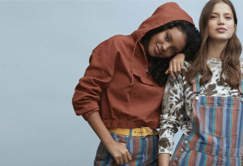 Two women posing in casual outfits; one wearing a rust-colored hoodie and striped bottoms, and the other wearing a tie-dye shirt with striped overalls.