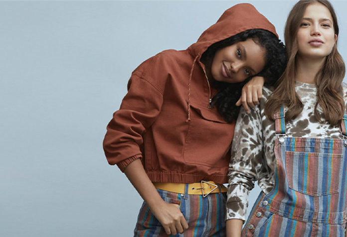 Two young women wearing casual outfits, one in a red hoodie and striped jeans, and the other in a tie-dye shirt with striped overalls.