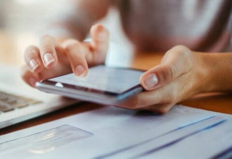 Person using a smartphone with documents and a laptop nearby.