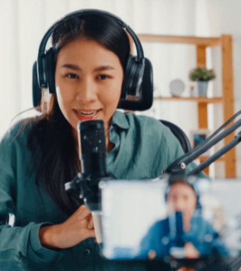 A woman wearing headphones speaks into a microphone, with a smartphone on a tripod recording her, in a room with shelves and natural light.