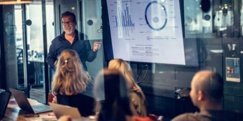 A person giving a presentation to a group in a meeting room, with graphs displayed on a large screen.