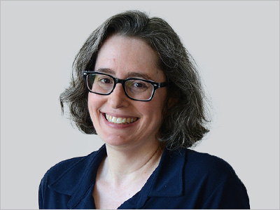 A person with shoulder-length hair and glasses, smiling, wearing a dark shirt.