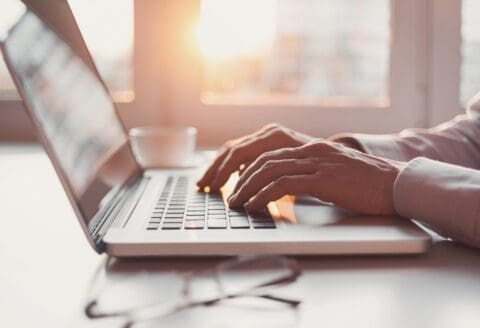 Hands typing on a laptop keyboard with a cup and glasses on a desk, illuminated by sunlight.