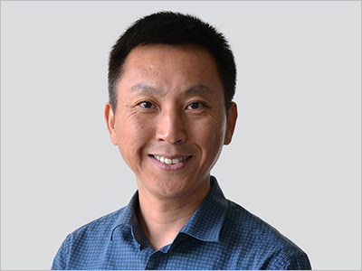 A man smiling, wearing a blue checkered shirt, with a gray background.