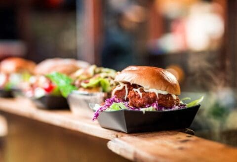 A close-up of a gourmet burger with toppings and sauce in a black tray on a wooden counter, with other burgers blurred in the background.