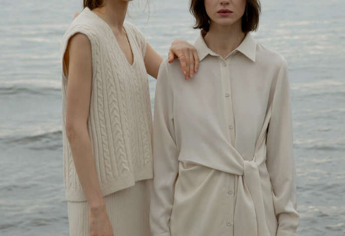 Two women wearing neutral-colored, minimalist clothing standing by the water.