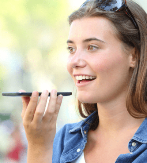 A woman holding a smartphone and speaking into it, appearing outdoors.