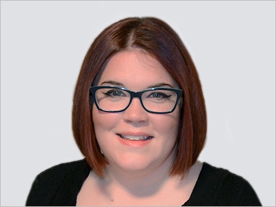 A person with red hair and glasses smiling against a gray background.