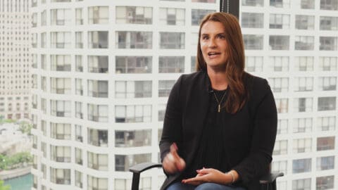 A woman sitting in an office chair speaking, with a background of large city buildings visible through the window.