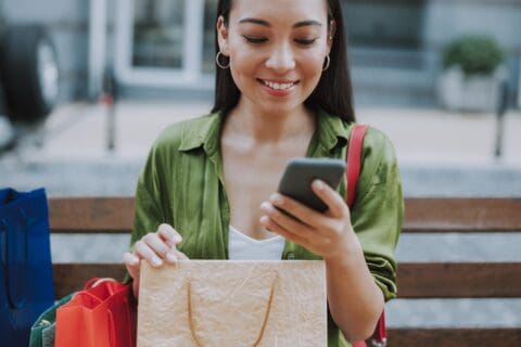 A woman sitting on a bench holding shopping bags and looking at her smartphone, smiling.