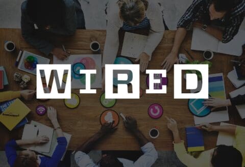 A group of diverse people sitting around a table with gadgets, papers, and coffee cups, with the word "WIRED" overlaid in bold white letters.