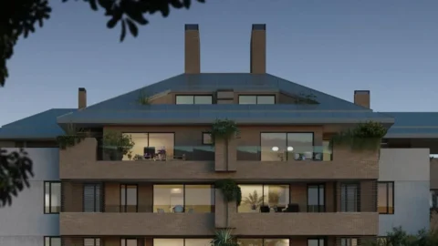 A modern multi-story brick building with large windows and balconies, surrounded by plants, seen at dusk.