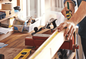 Person measuring a wooden plank with a tape measure in a workshop.