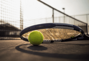 Tennis racket and ball on a court with a net in the background.