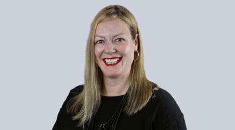 Smiling woman with blonde hair wearing a black top, against a light gray background.