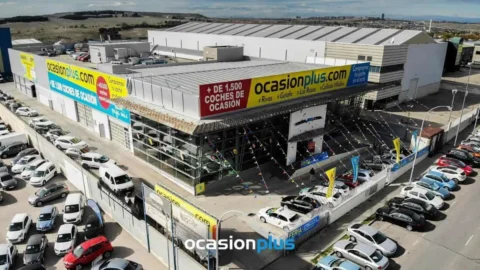 Aerial view of a large car dealership with vehicles parked outside, featuring prominent "ocasionplus.com" signage.