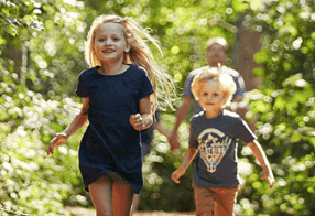 Children running joyfully through a forest path on a sunny day.