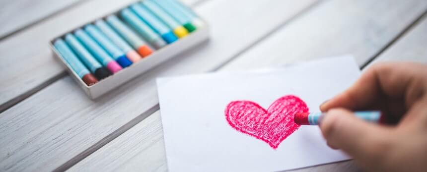 A hand drawing a red heart on paper with crayons nearby.