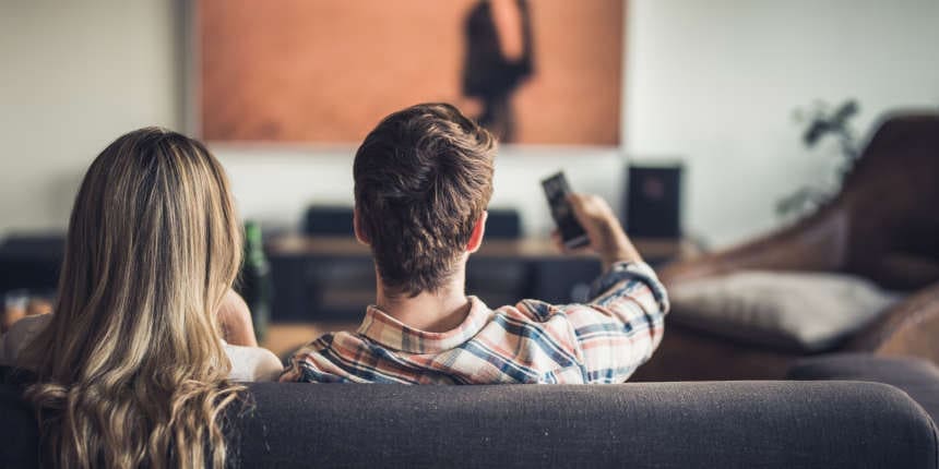 Back view of a couple watching TV on sofa at home. Man is changing channels.