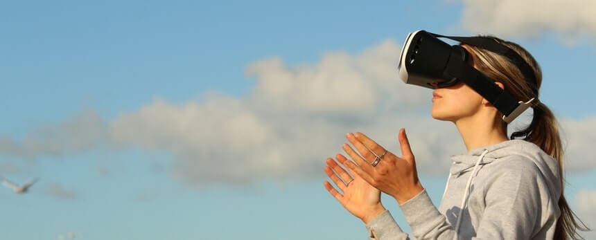 A woman wearing a virtual reality headset claps her hands against a blue sky with clouds.