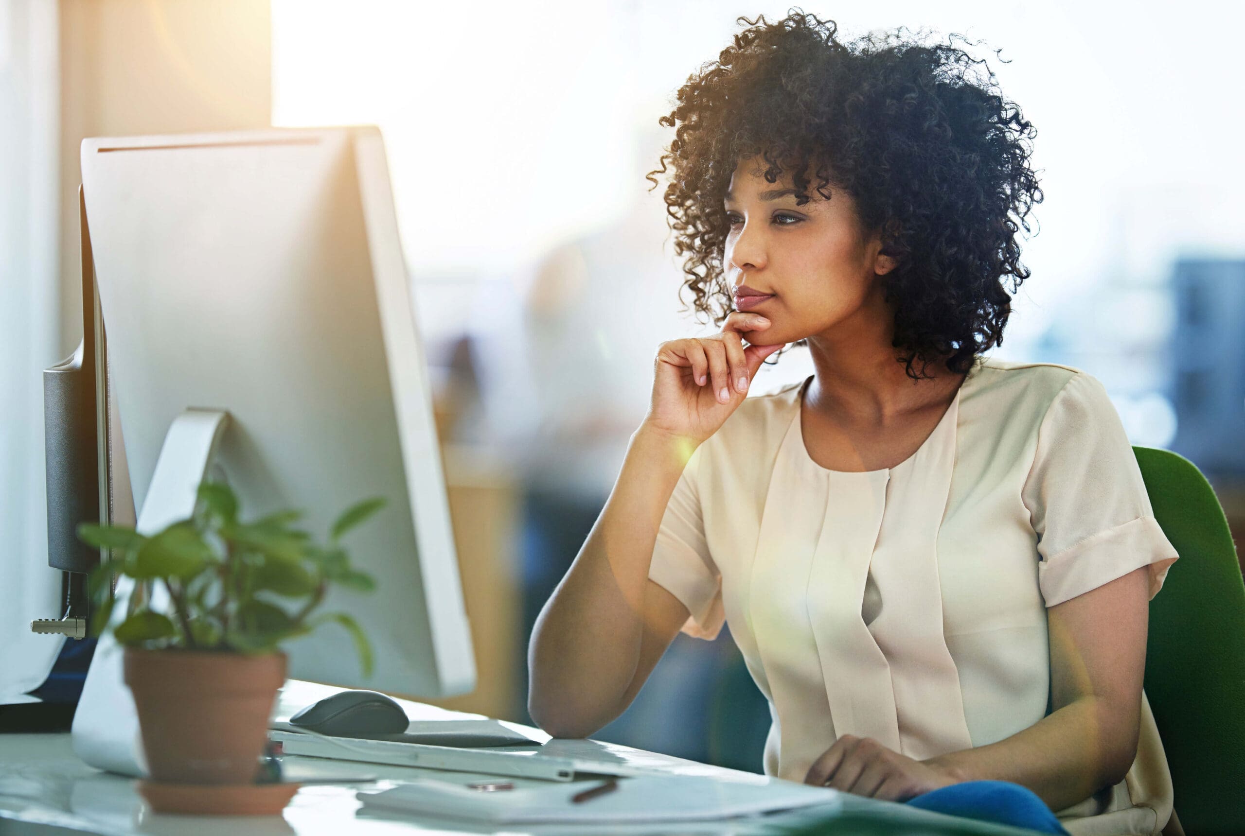 A woman sitting at a desk, looking thoughtfully at a computer screen, with a potted plant nearby.