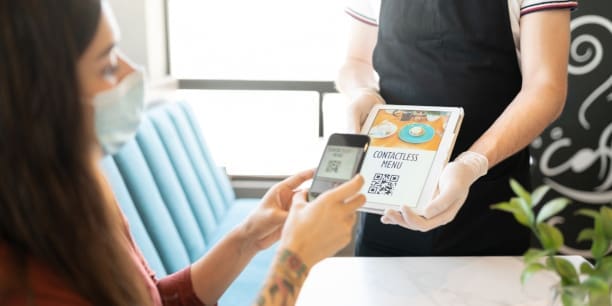 Young woman scanning QR code of contactless menu through smartphone in restaurant