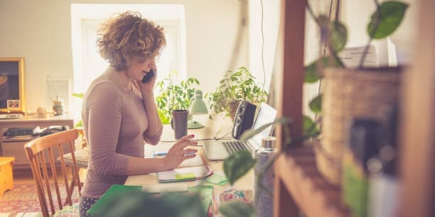 Woman working from home, using phone, sitting on the chair and writing into the book. 