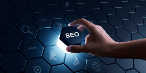 A hand holding a glowing hexagonal block labeled "SEO" against a dark hexagonal patterned background with various icons related to search and digital marketing.