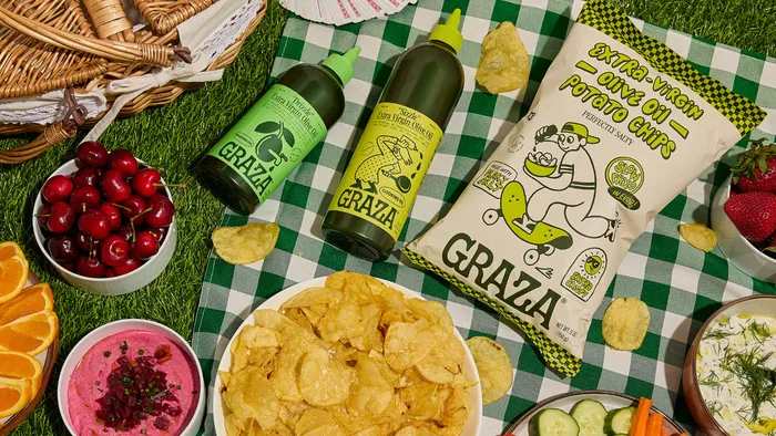 Picnic setup with a variety of snacks on a green checkered cloth, featuring Graza olive oil bottles, a large bowl of potato chips, a bag of Graza extra virgin olive oil potato chips, cherries, orange slices, and a bowl of pink dip.