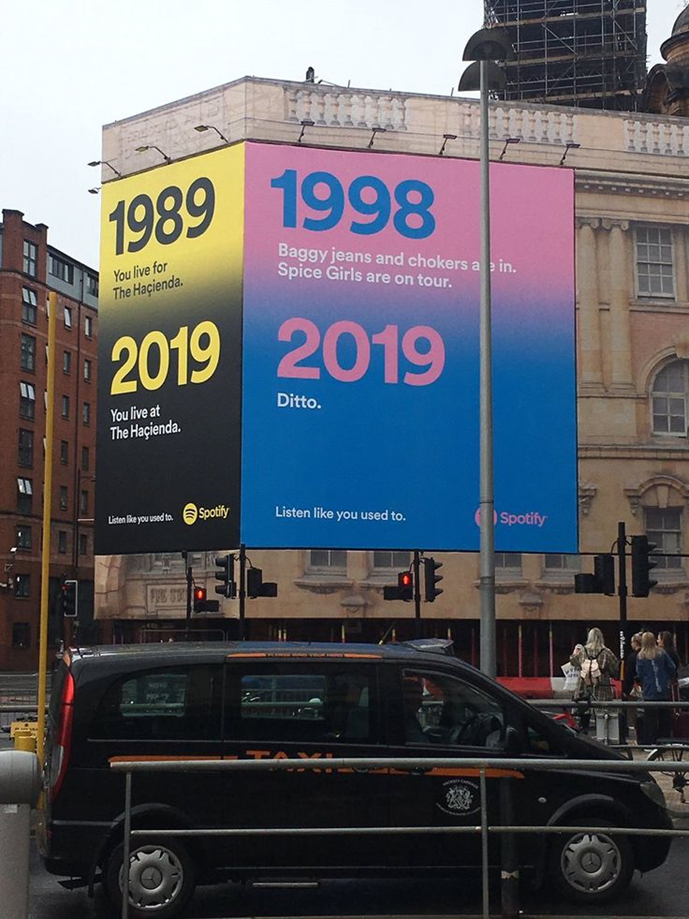 A two-part Spotify billboard featuring text emphasizing nostalgia, with the years 1989, 1998, and 2019, comparing cultural trends. A taxi is parked in front.